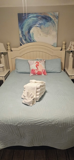 Un lit avec une couette bleue, un oreiller blanc orné d’un motif représentant un hippocampe, et une pile de serviettes blanches.