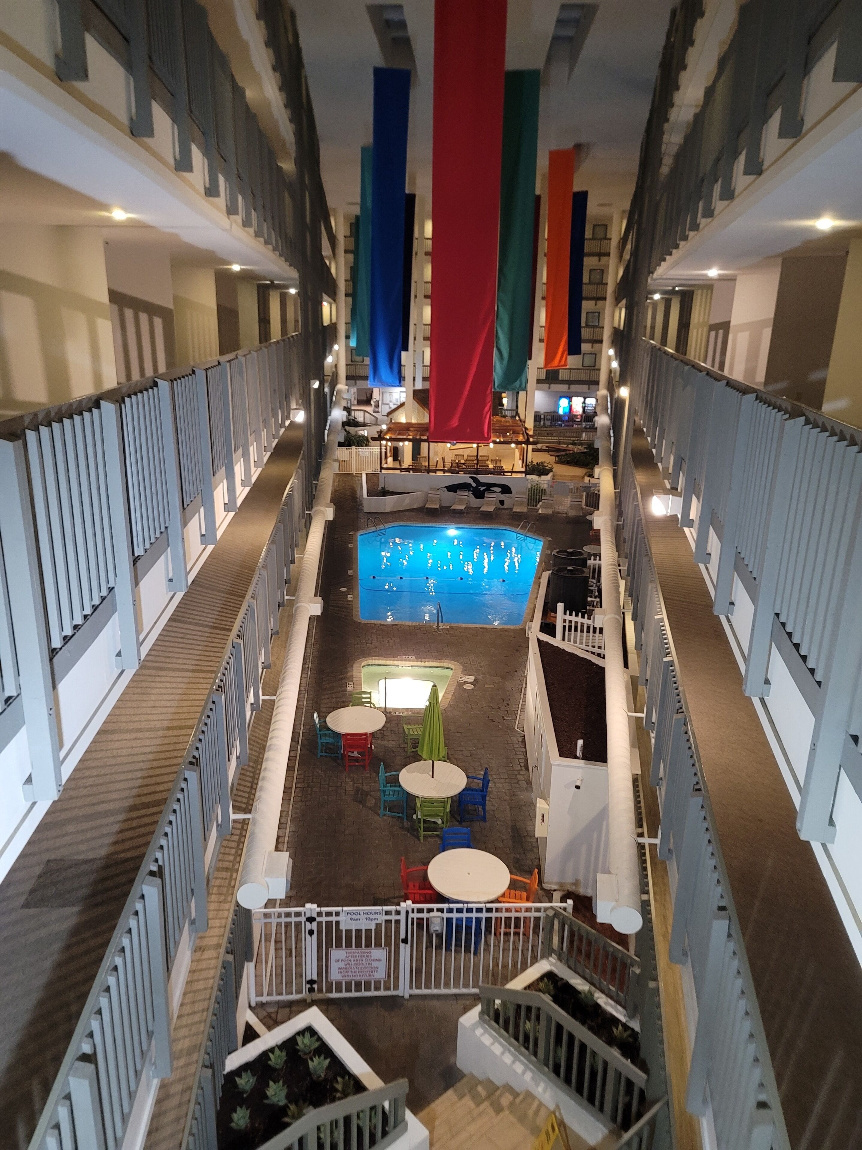 A pool area with a central pool, lounge chairs, and tables. There are colorful flags hanging from the ceiling.