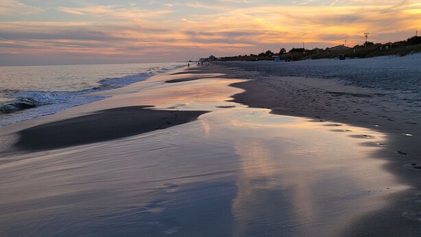 Ein Strand mit einer feuchten Sandfläche, die zum Wasser führt, einem klaren Himmel und Gebäuden in der Ferne.