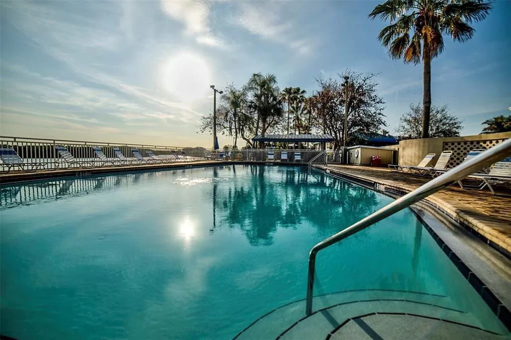 A clear swimming pool with lounge chairs and a covered area, surrounded by palm trees and a clear sky.