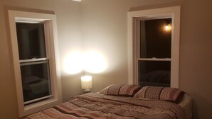 A bedroom with a bed, two windows, a nightstand, and a wall-mounted lamp. - Fully renovated luxury waterfront cottage (Havelock)