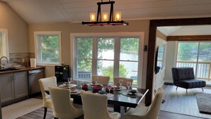 A dining area with a table set for four, surrounded by chairs, with a view of a lake through large windows. - Fully renovated luxury waterfront cottage (Havelock)