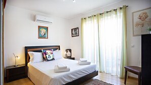 A bedroom with a large bed, a nightstand with a lamp, an air conditioner, and a window with curtains. - Cabanas Gardens,  6px, terrace and barbecue (Cabanas de tavira)