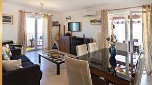 A modern living room with a glass dining table, a sofa, a TV, and a guitar. - Cabanas Gardens,  6px, terrace and barbecue (Cabanas de tavira)
