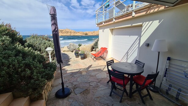 A patio with a table and chairs, a covered umbrella, and a view of the sea and a lighthouse in the background. - Self-contained 75 m2 apartment on the ground floor of a villa, air-conditioned, seafront (L'Île-Rousse)