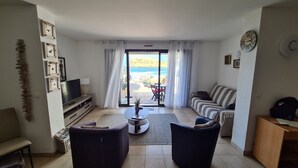 A living room with a TV, sofa, and chairs, a view of the sea through a glass door, and a clock on the wall. - Self-contained 75 m2 apartment on the ground floor of a villa, air-conditioned, seafront (L'Île-Rousse)