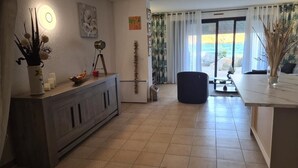 A modern living room with a dining area, a large window with curtains, and a view of the sea. - Self-contained 75 m2 apartment on the ground floor of a villa, air-conditioned, seafront (L'Île-Rousse)
