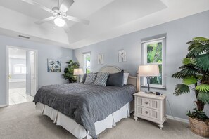 Un dormitorio con cama, mesitas de noche, ventilador de techo, ventana con persianas y una planta en maceta.