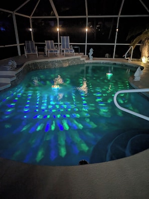 Una piscina con efectos de iluminación azul, rodeada de sillones de descanso y una zona cubierta con escalones que dan a la piscina.