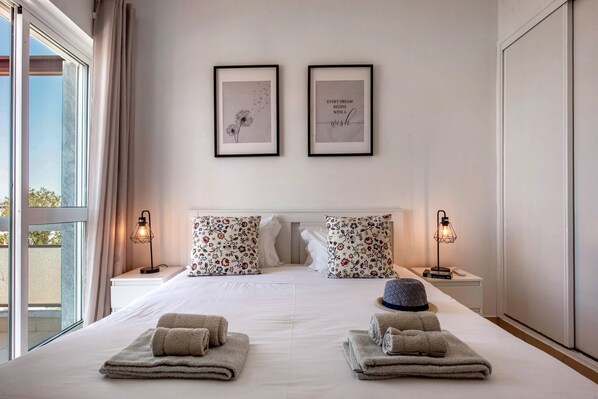 A hotel room with a large bed, two framed pictures on the wall, a window with curtains, and a nightstand with a lamp. - Apartment in the city center! (Faro)