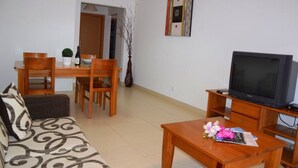 A living room with a sofa, a coffee table with flowers, a TV on a wooden stand, a dining area with chairs and a table, and a painting on the wall.