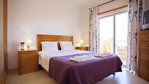 A hotel room with a wooden bed, two bedside tables with lamps, a purple bedspread, and a view of the outside through a sliding glass door.
