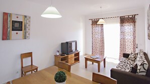 A living room with a wooden dining table, a sofa, a TV on a wooden stand, and a potted plant.