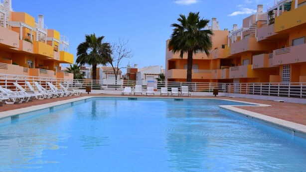 A swimming pool with white lounge chairs and palm trees, surrounded by multi-story buildings with balconies.