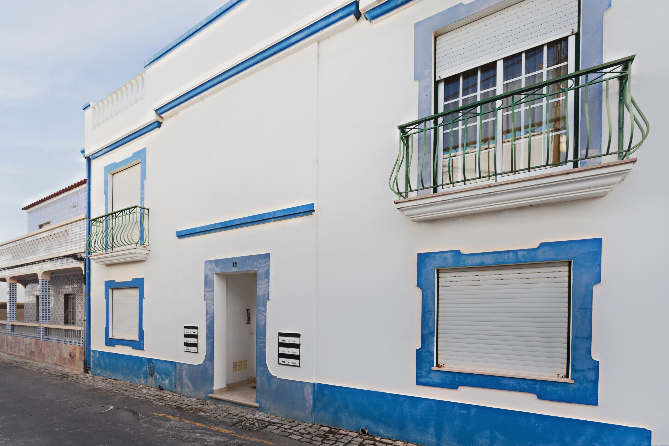 A building with a white facade, blue trim, a balcony with green railings, and a closed shuttered window.