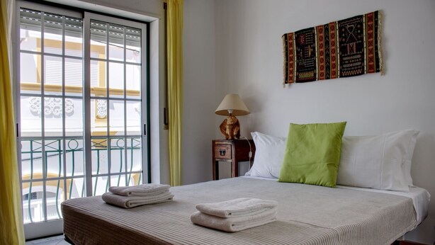 A hotel room with a bed, a nightstand with a lamp, a window with blinds, and a balcony view.