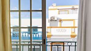 A view through a window with a metal grid, a balcony with a blue railing, and a white building with yellow trim and a clock.