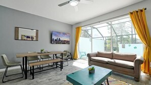 A modern living room with a sofa, a dining table with chairs, a coffee table, and a large window with curtains. - Windsor Hills Home w/ Pool 2 miles from Disney! (Kissimmee)