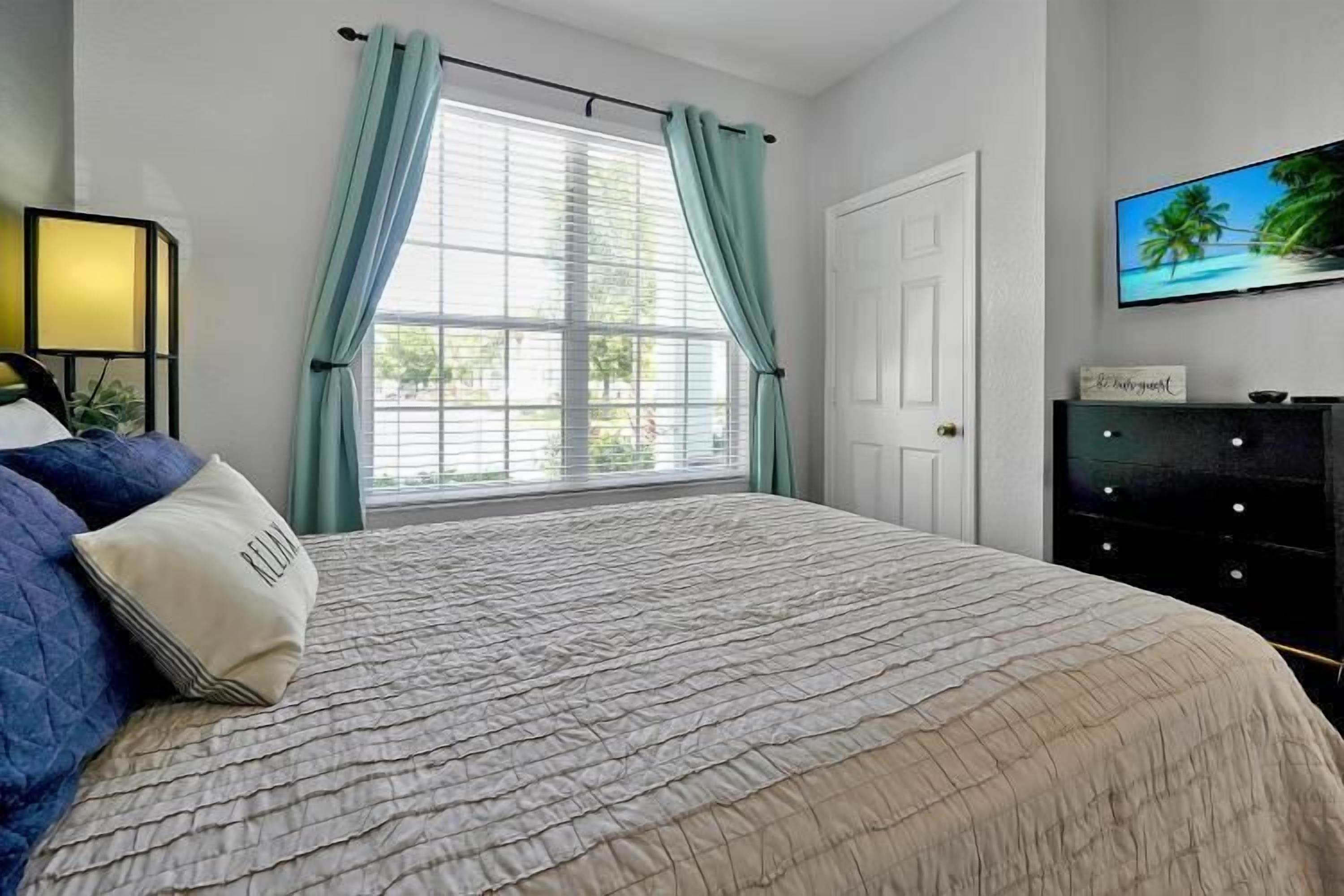 Une chambre à coucher avec un grand lit, une fenêtre avec des rideaux, un téléviseur fixé au mur et une commode.