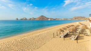 A beach with rows of sun loungers and umbrellas, a clear blue sky, and distant mountains. - Located on the pristine Medano beach (Cabo San Lucas)