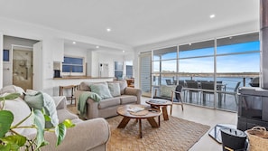 A spacious living area with a large window overlooking a body of water, a kitchen with a view, and a balcony with outdoor seating.