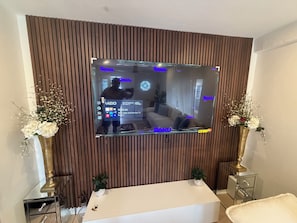 A modern living room with a large flat-screen TV mounted on a wooden wall, a white sofa, and two decorative vases with flowers. - Walk to Beach 2- Patios Updated Lux Retreat NEW Heated Pool Sleeps 10 Small Dogs (Naples)
