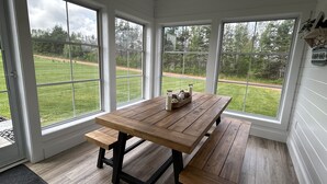 Een kamer met een houten tafel en bankjes, grote ramen met uitzicht op een grasveld en bomen.