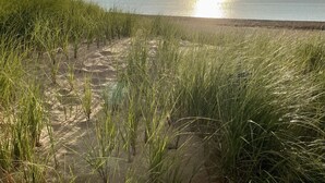 Een strand met zand en hoge grassen op de voorgrond, een waterpartij in het midden en een ondergaande zon op de achtergrond.