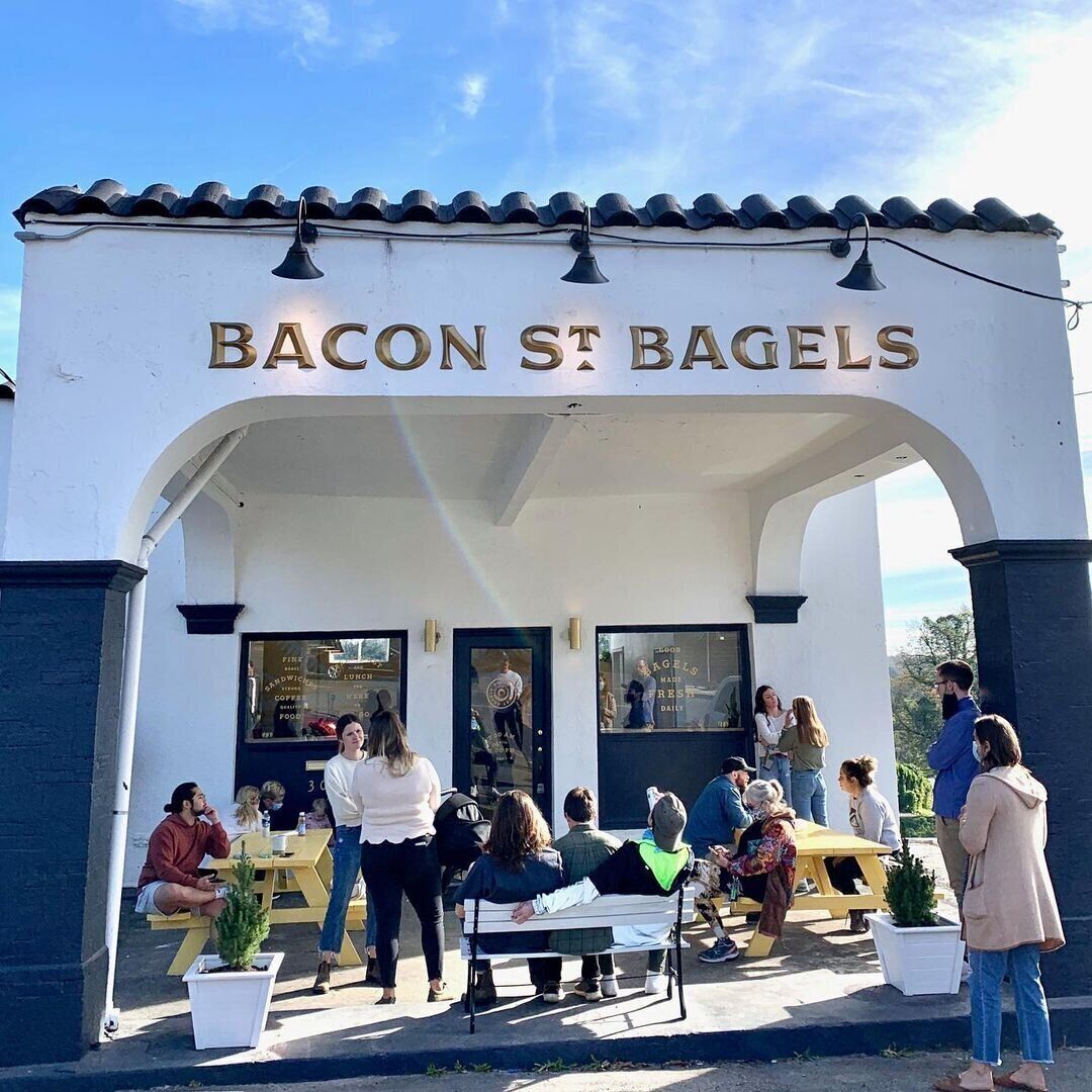 Fasaden på en restaurang som heter ”Bacon St Bagels”, med uteservering och en klar himmel.