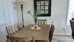 Une salle à manger avec une table et des chaises en bois, un vase avec une plante, une fenêtre avec des stores et une lampe.