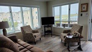 Un salon avec un canapé marron, des fauteuils à motifs, un meuble pour la télévision et une table sur laquelle se trouve un bol de collations. De grandes fenêtres offrent une vue sur un quartier résidentiel.