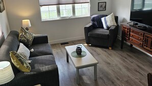 Een woonkamer met een bank, fauteuil, salontafel, televisie en een raam met jaloezieën.