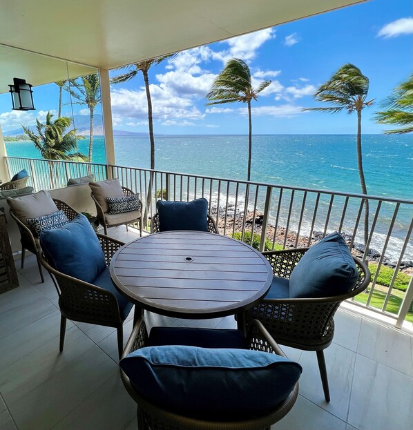 A balcony with wicker furniture, a round table, and a view of the ocean and palm trees.