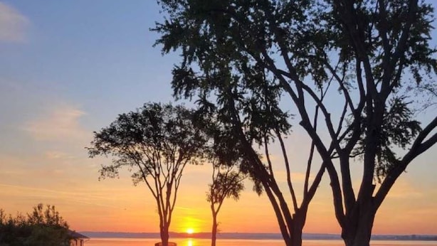 A sunset view from a deck overlooking a grassy area with trees and a body of water. - Beautiful Sunrises and Lakefront Views! (Fond du Lac)