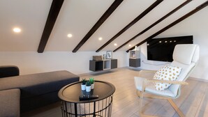 Quarto moderno estilo loft com teto inclinado, uma cama grande, área de estar com sofá e poltrona, uma pequena mesa redonda com uma planta e prateleiras embutidas.