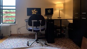 A room with a desk, chair, computer, lamp, radiator, and a window with a view of greenery. - True 1Bdrm (New York)