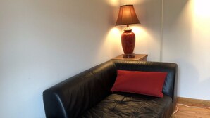 A black leather sofa with a red cushion in a room with wooden flooring and a lamp on a wooden side table. - True 1Bdrm (New York)