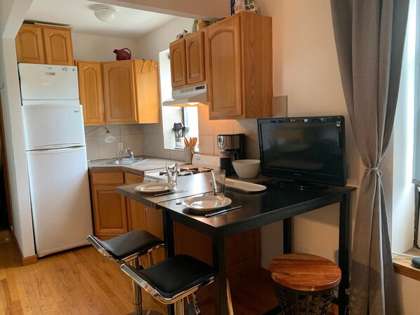 A compact kitchen with wooden cabinets, a white refrigerator, a black countertop, and a small dining table with two chairs.