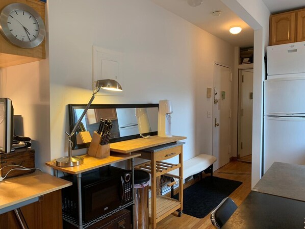 A compact kitchen with a microwave, stove, and a wooden countertop. There is a clock on the wall and a refrigerator in the background.