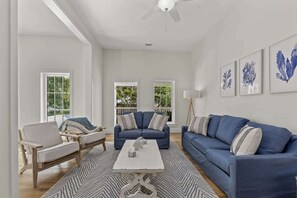 A spacious living room with a blue sofa, a white coffee table, and a patterned rug. There are two windows with white frames and a ceiling fan. - Luxury 8BR Home w/ Pickleball, Pool & Spa (Santa Rosa Beach)