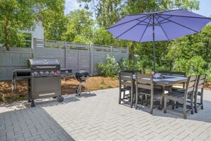 Um pátio externo com churrasqueira, mesa e cadeiras, e um guarda-sol roxo.