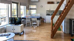 Een ruime woonkamer met een houten vloer, een eettafel met stoelen, een televisie op een standaard, en een keuken met kastjes en een klok.