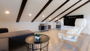 Ein modernes Schlafzimmer im Loft-Stil mit schrägem Dach, einem großen Bett, einer Sitzecke mit Sofa und Sessel, einem kleinen runden Tisch mit Fernbedienung und einem an der Wand montierten Fernseher.