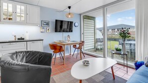A modern kitchen with white cabinets, a dining area with a white table and orange chairs, and a large sliding glass door leading to an outdoor patio with a view of houses. - 1 bedroom beautiful home in Fanø (Fanø)