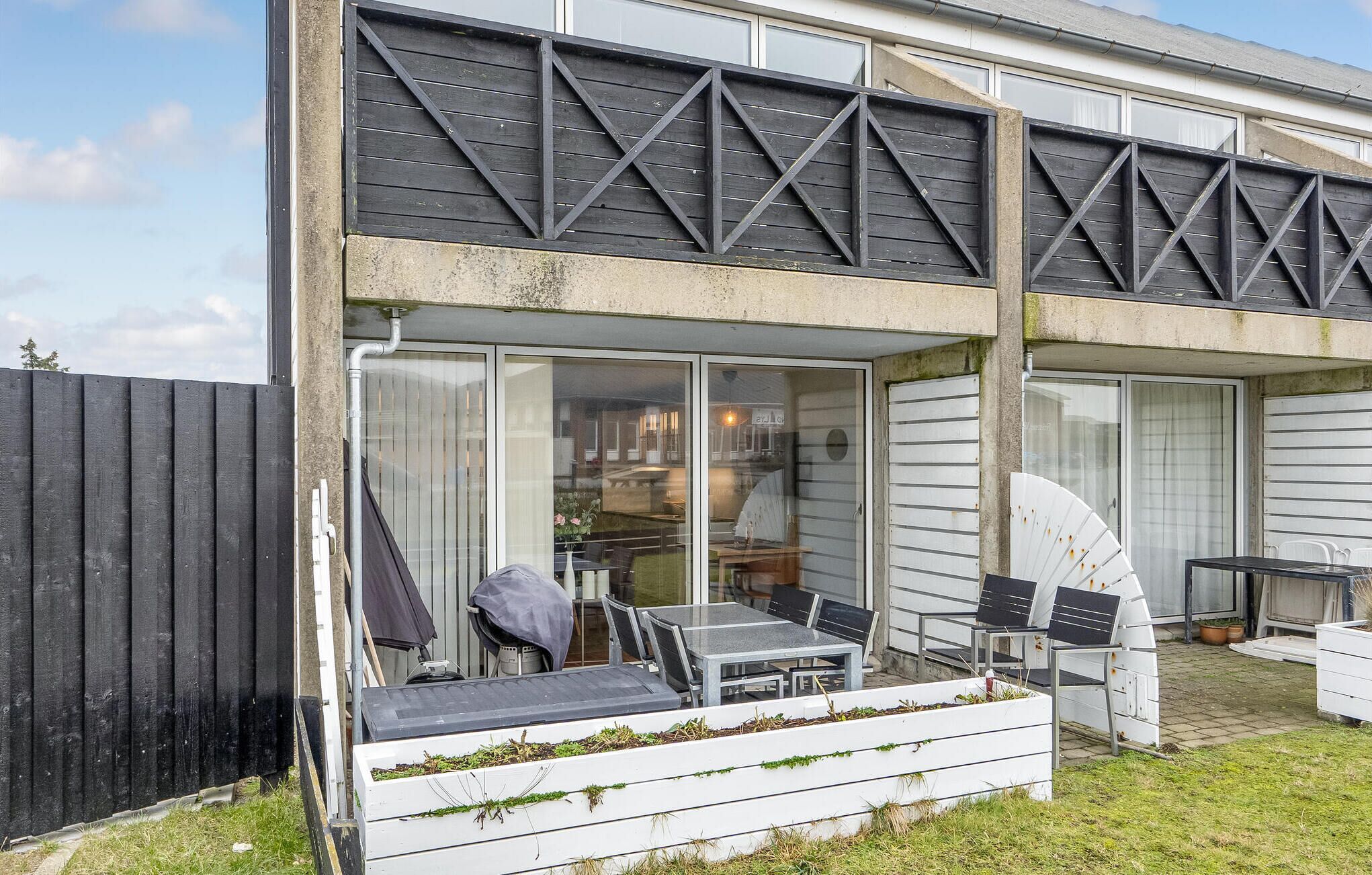 Une maison moderne dotée d’un patio comprenant une table et des chaises, un barbecue et un bac à fleurs blanc orné de plantes vertes.