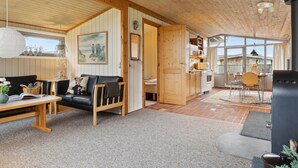 A cozy living room with a sofa, a coffee table, and a painting on the wall. There is a wooden door leading to a kitchen area with a dining table and chairs.