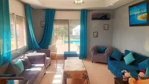 A living room with a sofa, armchair, and coffee table, a large window with curtains, and a painting on the wall. - American Farm Villa El Jadida, Sidi Rahal, Azemour  (Casablanca-Settat)
