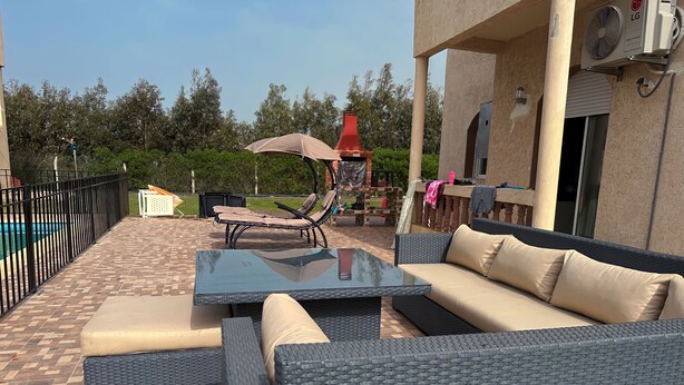 An outdoor patio with wicker furniture, a glass table, and a pool in the background. - American Farm Villa El Jadida, Sidi Rahal, Azemour  (Casablanca-Settat)