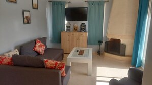 A living room with a sofa, a coffee table, and a television. There are curtains and a cabinet with decorative items. - American Farm Villa El Jadida, Sidi Rahal, Azemour  (Casablanca-Settat)
