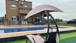 A poolside area with a swing chair and a covered umbrella, a pool, and a building with a balcony in the background. - American Farm Villa El Jadida, Sidi Rahal, Azemour  (Casablanca-Settat)